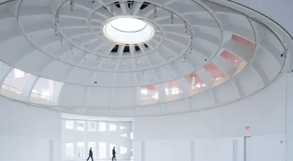 The expansive, white interior of the Faena Art Project Room features a striking domed ceiling with a central skylight. Two figures walk through the minimalist space.
