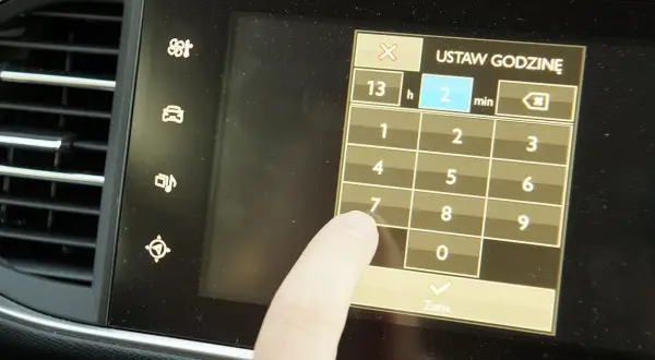 Palec dotyka ekranu podczas ustawiania godziny w Opel Astra H. Wyświetlacz pokazuje klawiaturę numeryczną do wprowadzania czasu.