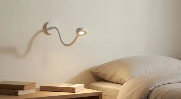 Biała, elastyczna lampa nocna oświetla stos książek na stoliku obok łóżka.
