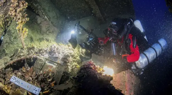 Nurkowie badają wrak statku, być może **Britannic**, z kamerą i oświetleniem. Widoczne są szczątki, muszle i ciemne głębiny.