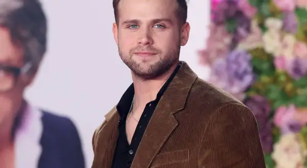 Leo Woodall, con su chaqueta de pana marrón y camisa negra, sonríe ante la cámara.