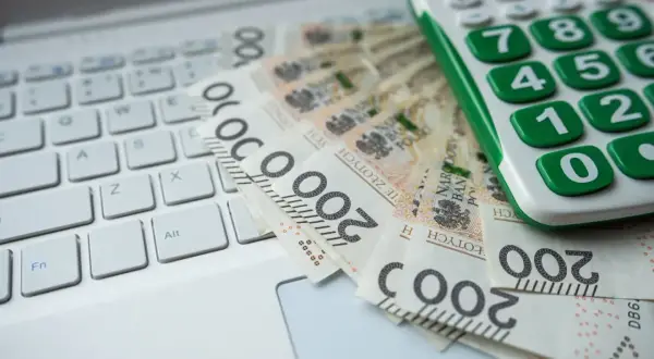 Kalkulator i banknoty 200 zł na klawiaturze laptopa. Obrazek sugeruje analizę finansową, być może związaną z grupami podatkowymi.