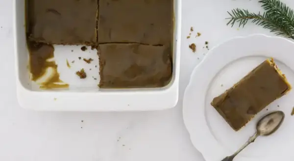 En bit av en saftig sticky toffee pudding med kolaglasyr på en vit tallrik, bredvid en form med resten av kakan.