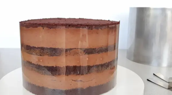 Czekoladowa torta z warstwami ciasta i kremu, przygotowywana w transparentnej formie. Obok stoi metalowy pierścień.