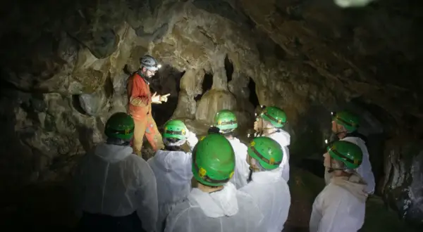 Exploradores en la cueva Rei Cintolo, con cascos verdes y trajes blancos, escuchan atentamente a su guía.
