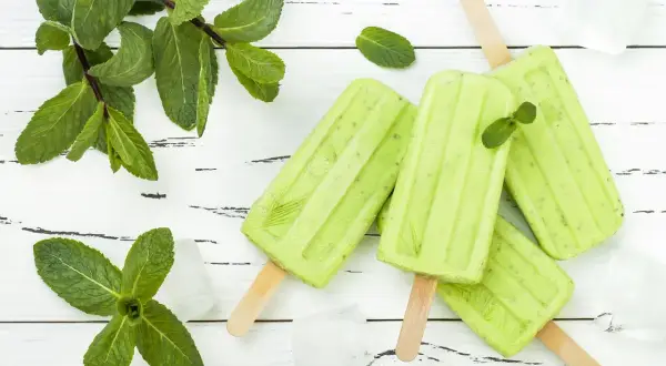 Erfrischendes Minzeis selber machen: Grüne Eis am Stiel mit frischer Minze und Eiswürfeln auf weißem Holzuntergrund.