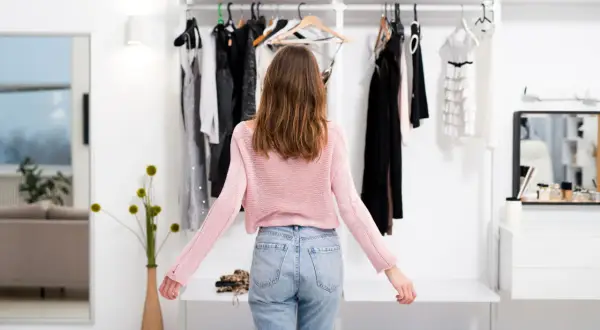 Dziewczyna w różowym swetrze i jeansach robi outfit check przed szafą pełną ubrań.