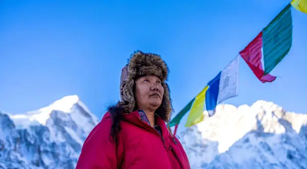 Lhakpa Sherpa, eine Frau mit einem Pelzmützen, blickt in den Himmel, während Gebetsfahnen über schneebedeckten Bergen wehen.