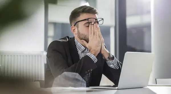 Man med glasögon sitter vid en laptop, gnuggar ögonen. Ett klassiskt dilemma exempel på stressad arbetsdag.
