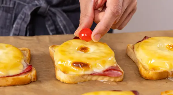 Eine Hand platziert eine Kirsche auf einem überbackenen Toast Hawaii. Der Toast im Backofen ist mit Schinken, Ananas und geschmolzenem Käse belegt.