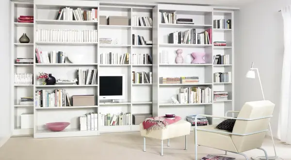 Eine moderne, weiße **eigene Bibliothek** mit vielen Büchern, Dekorationen und einem Fernseher. Ein Sessel und ein Hocker laden zum Verweilen ein.