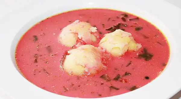 Pyszna zupa z botwiny z ziemniakami, podana w białej misce. Trzy kluski ziemniaczane unoszą się na różowym, aromatycznym bulionie.