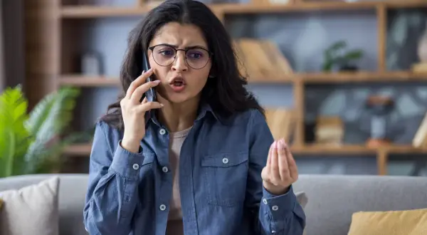 Donna con occhiali parla al telefono con espressione frustrata, un gesto della mano che suggerisce il desiderio di chiudere il telefono in faccia psicologia.