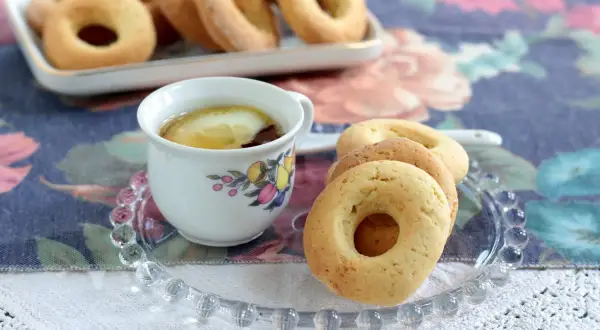 Biscotti senza burro dorati su un piattino di vetro, accanto a una tazza di tè con limone.