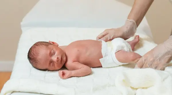 Noworodek z hipotrofią płodu leży na białym kocyku, a ręce w rękawiczkach delikatnie go dotykają.