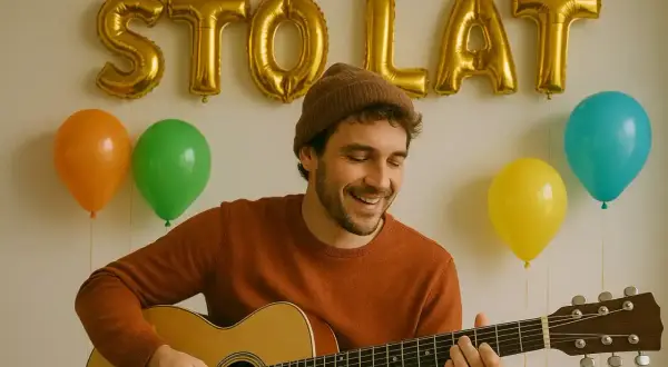 Uśmiechnięty mężczyzna gra na gitarze, a za nim złote balony tworzą napis "STO LAT". Dowiedz się, jak zagrać sto lat na gitarze.