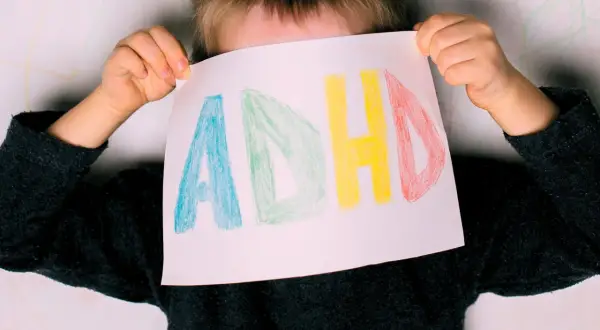 Dziecko z ADHD trzyma kartkę z napisem "ADHD" narysowanym kolorowymi kredkami.