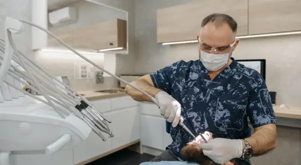 Dentysta w masce i okularach wykonuje zabieg, być może kortykotomię, u pacjenta leżącego na fotelu.