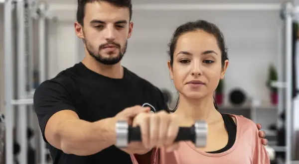 Trener pomaga kobiecie ćwiczyć z hantlem. To przykład, jak fitness co to jest – aktywność fizyczna dla zdrowia i lepszego samopoczucia.