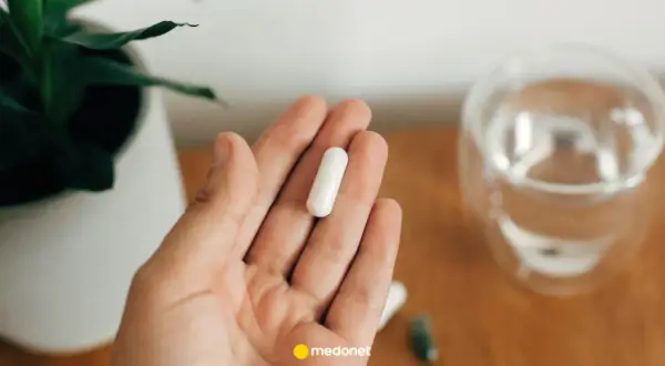 Dłoń trzyma białą kapsułkę. Obok szklanka wody i roślina. Uważaj na nadmiar magnezu objawy.