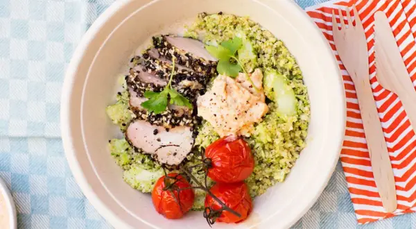 En skål med tillagad quinoa, skivat kött med sesamfrön, hummus, gurka och körsbärstomater. Perfekt för en snabb och hälsosam lunch.