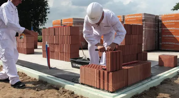 Dwóch budowlańców w białych kombinezonach i kaskach układa cegły na fundamencie.