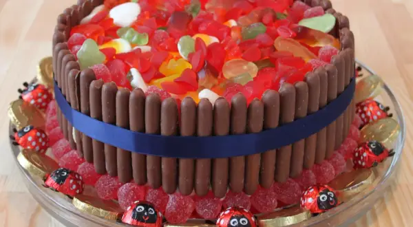 En tårta dekorerad med godis, chokladpinnar och en blå sidenband. Runt tårtan finns godis som ser ut som nyckelpigor och guldfärgade chokladmynt.