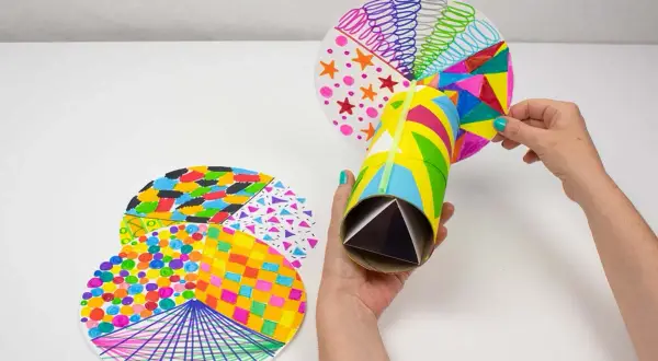 Manos creando un caleidoscopio con manualidades con material reciclado. Tubo de cartón decorado y discos circulares con patrones coloridos.