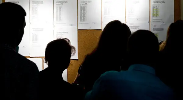 Sylwetki ludzi patrzą na tablicę ogłoszeń z listami wyników rekrutacji do szkół.