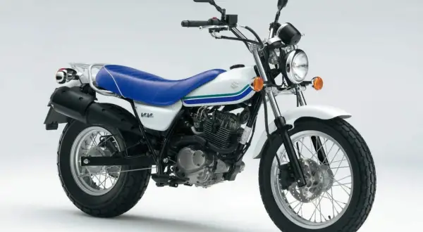 Biały motocykl Suzuki Van Van z niebieskim siedzeniem i zielono-niebieskimi pasami na baku. Idealny na miejskie wyprawy.