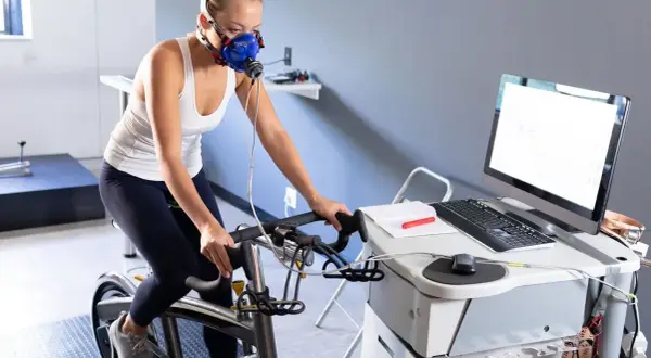 Femme en masque respiratoire effectuant un test d'effort duree sur un vélo d'appartement, connectée à un équipement médical.