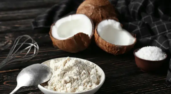 Miska pełna mąki kokosowej, obok połówki kokosa, wiórki kokosowe i trzepaczka.