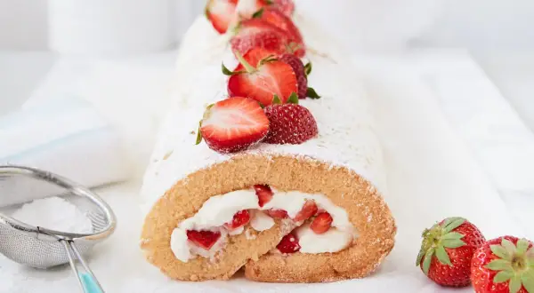 Saftige erdbeer biskuitrolle mit frischen Erdbeeren und Puderzucker bestäubt, daneben ein Sieb mit Puderzucker.