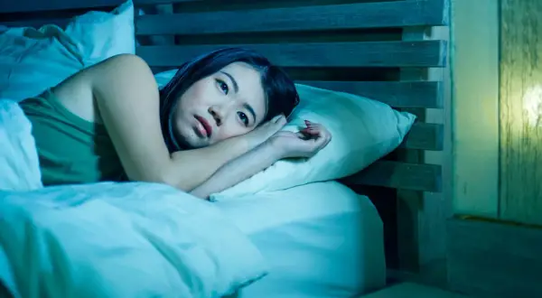 A woman lies awake in bed, her eyes wide open, a picture of paradoxical insomnia. The room is bathed in a cool, blue light, emphasizing her distress.