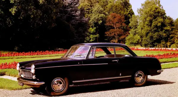 Elegancki, czarny Peugeot 404 coupe z lat 60. stoi na żwirowej alejce, otoczony zielenią i czerwonymi kwiatami.