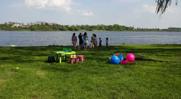 Dzieci bawią się nad jeziorem Mikorzyńskim. Na trawie kolorowe zabawki: piłki, stolik i krzesełka.