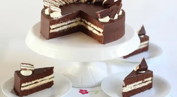 Schokoladentorte mit cremiger Füllung und Schokostückchen, inspiriert von der Kinder Pingui Torte. Serviert auf einem Tortenständer mit einzelnen Tortenstücken.