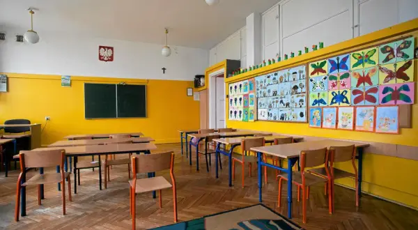 Pusta klasa szkolna z żółtymi ścianami, ławkami, krzesłami i kolorowymi pracami dzieci.