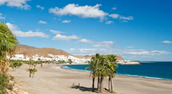La **playa de la Galera** se extiende bajo un cielo azul con nubes blancas. Palmeras salpican la arena dorada, mientras el mar turquesa baña la costa. Al fondo, un pueblo blanco se asoma entre las colinas.