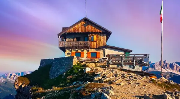 Rifugio Nuvolau thront auf einem Felsvorsprung unter einem lila Himmel. Eine italienische Flagge weht im Wind.