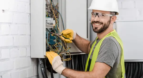 Uśmiechnięty technik elektryk w białym kasku i żółtych rękawicach pracuje przy skrzynce z przewodami.