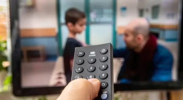 Dłoń trzymająca pilot do telewizora, na ekranie widać scenę z dzieckiem i mężczyzną. Czas na ulubiony serial, opłacony abonament radiowo-telewizyjny działa!