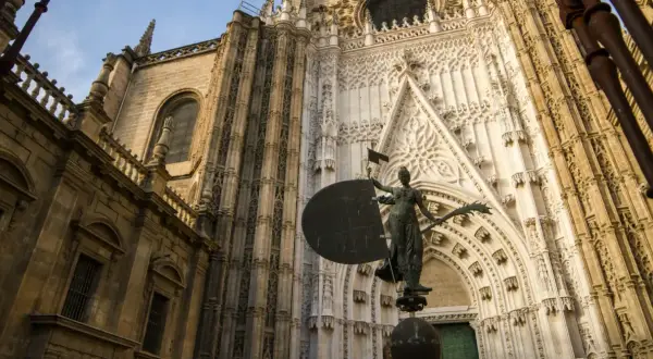 Estatua frente a la imponente fachada de la catedral de Sevilla, con detalles góticos y una escultura que evoca la navegación.
