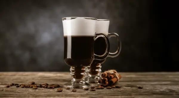 Två glas med kaffe med glass och vispgrädde, omgivna av kaffebönor och kanelstänger på ett träbord.
