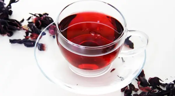 Szklana filiżanka z czerwonym naparem, obok suszone płatki hibiskusa. Idealny napój na odchudzanie.