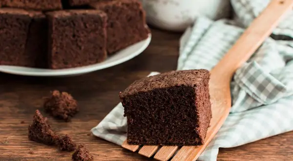 Un trozo de bizcocho de chocolate esponjoso servido en una espátula de madera, con más trozos apilados en un plato al fondo.