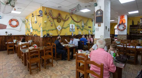 Gente comiendo en la abacería el arrecife, decorada con motivos marineros. Mesas de madera y paredes amarillas con redes y barcos.