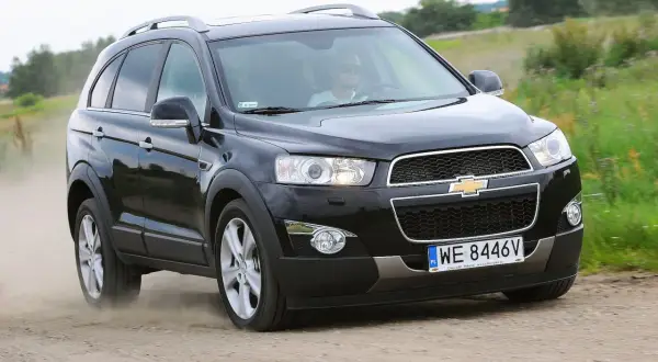 Czarny Chevrolet Captiva jedzie po piaszczystej drodze, wzbijając tumany kurzu. Kierowca w okularach przeciwsłonecznych prowadzi auto przez zielone pola.