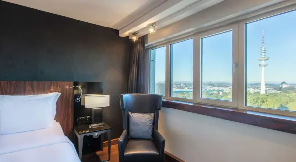 Pokój w Blu Radisson Hamburg z widokiem na wieżę telewizyjną. Wygodne łóżko, fotel i stolik nocny.