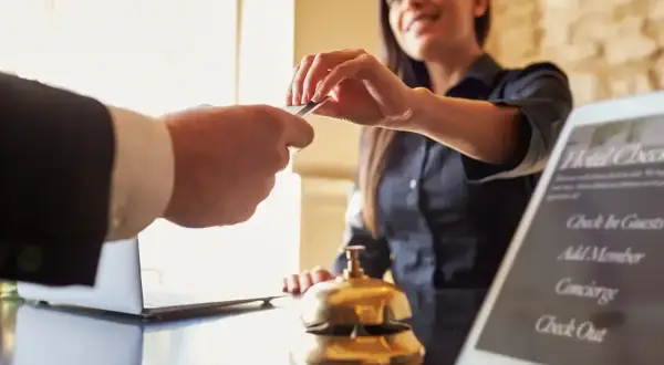Recepcjonistka uśmiecha się, przyjmując kartę od gościa. Na ekranie tabletu widoczna jest opcja "Check Out".